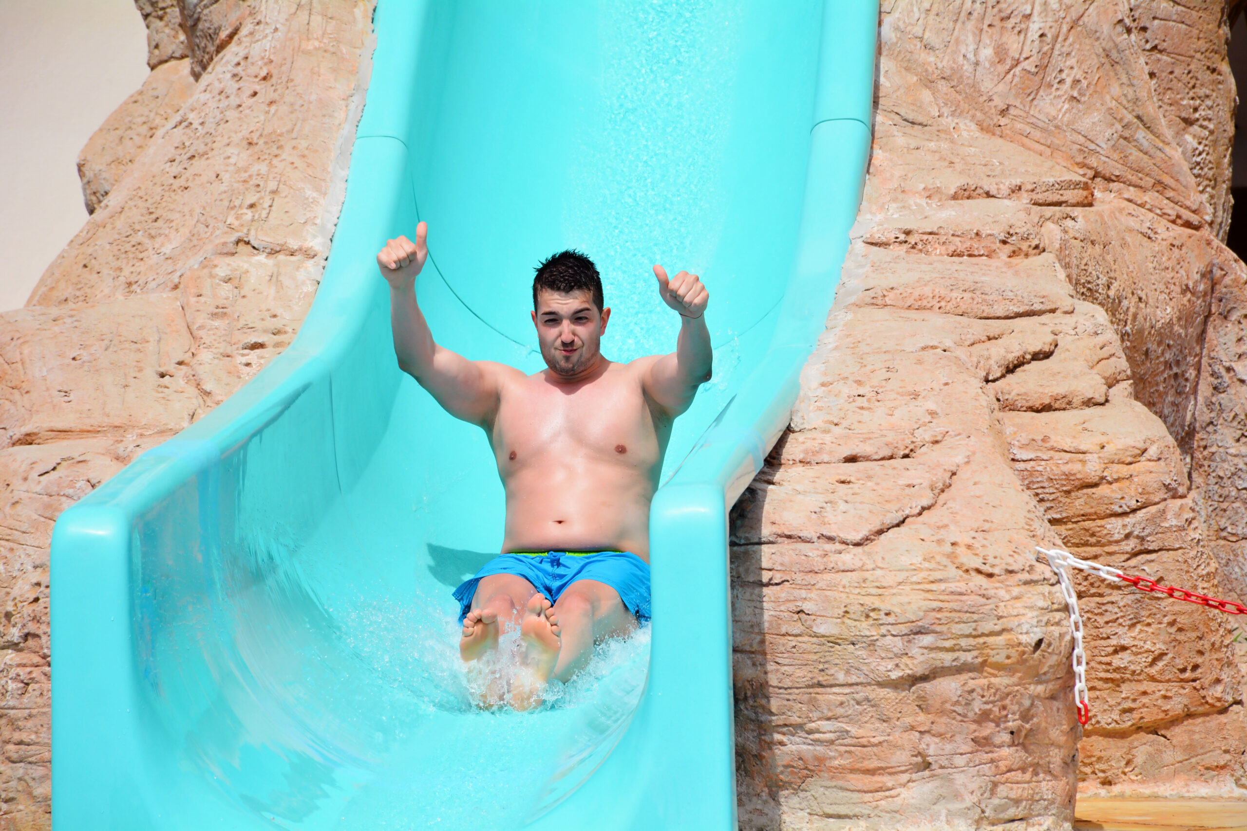 Hombre feliz haciendo la señal de pulgar arriba mientras se desliza por un tobogán de agua azul brillante en un parque acuático o resort