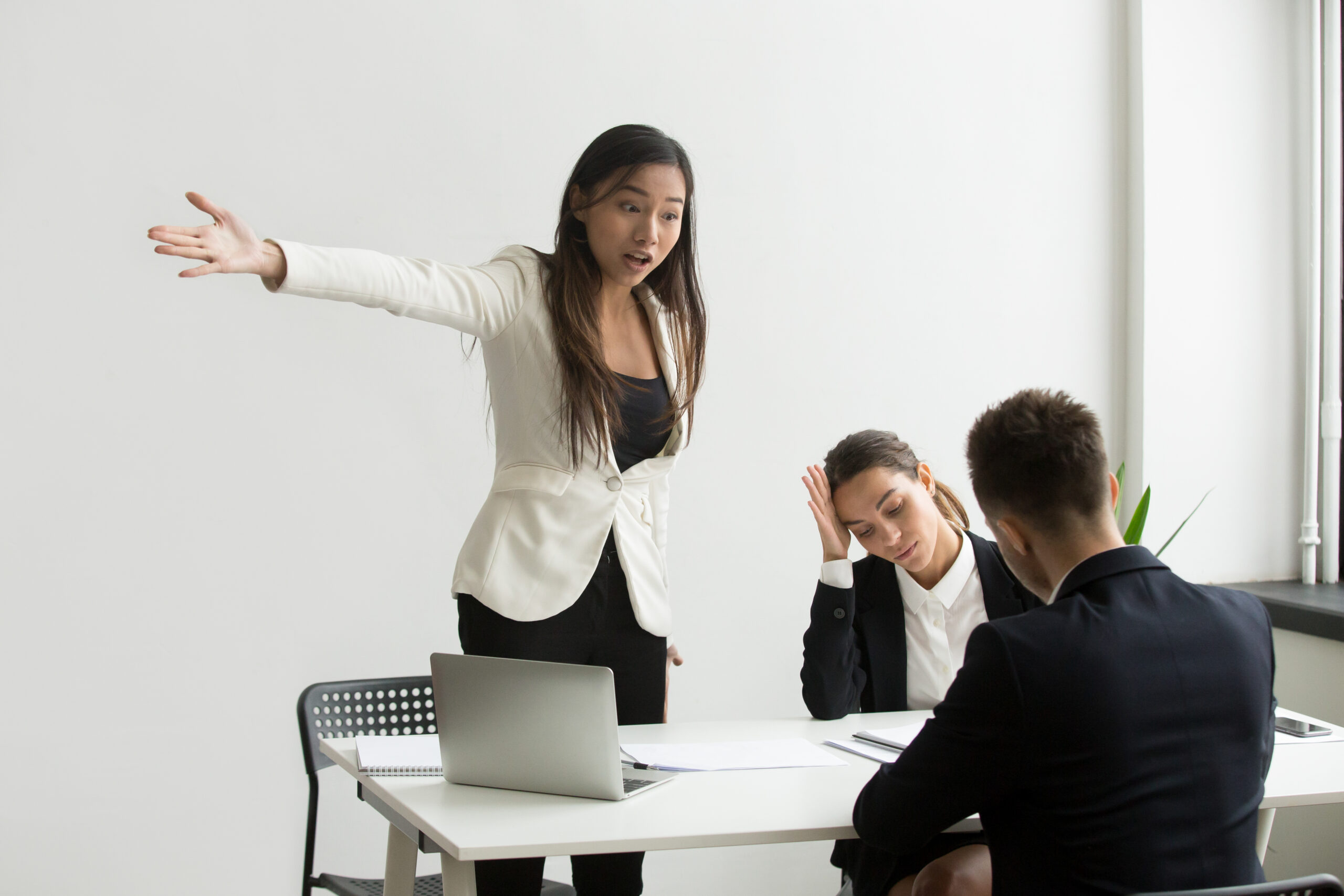 Gerente femenina enojada gritando y gesticulando a dos empleados que lucen estresados durante una tensa reunión en una oficina blanca y austera