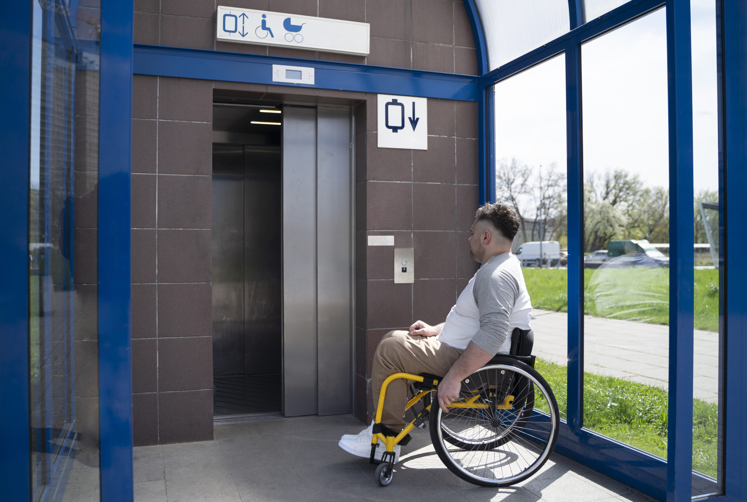 Un hombre en una silla de ruedas manual esperando un elevador público al aire libre con señalización de accesibilidad