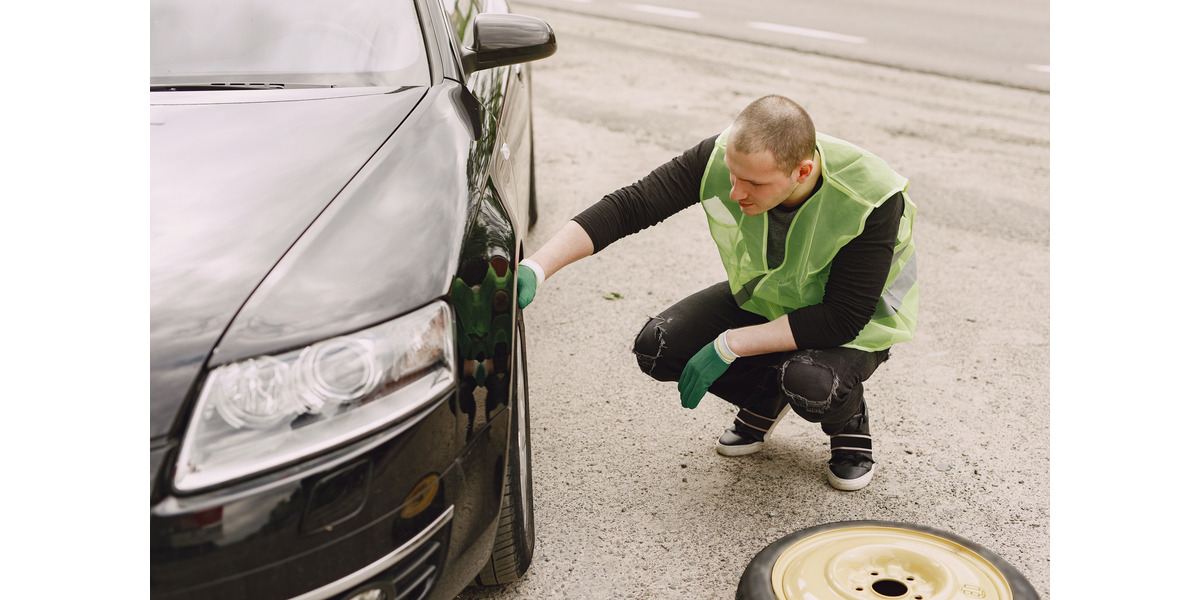 Hombre con chaleco de seguridad cambiando una llanta pinchada en un auto negro al costado de la carretera