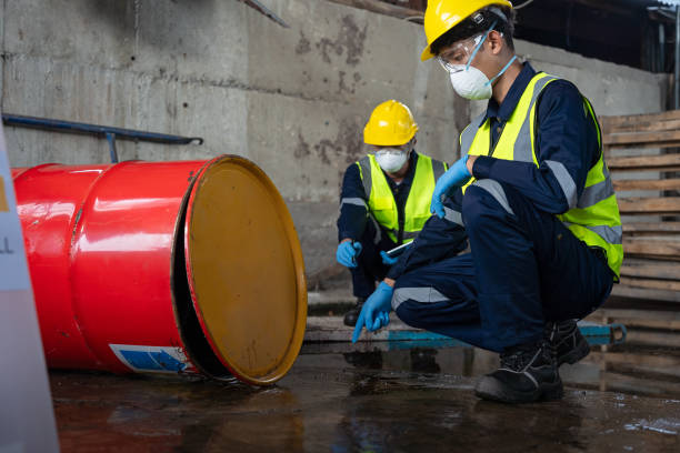 Dos trabajadores con chalecos de seguridad, cascos y respiradores inspeccionando un derrame químico de un tambor rojo volcado