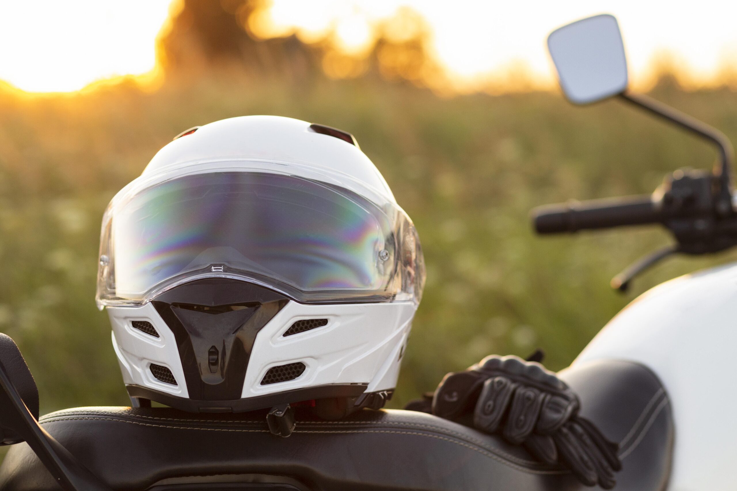 Casco blanco de motocicleta con visor reflectante sobre el asiento de una moto al atardecer, con guantes de conducción cercanos, representando equipo de seguridad y estilo de vida de motociclismo
