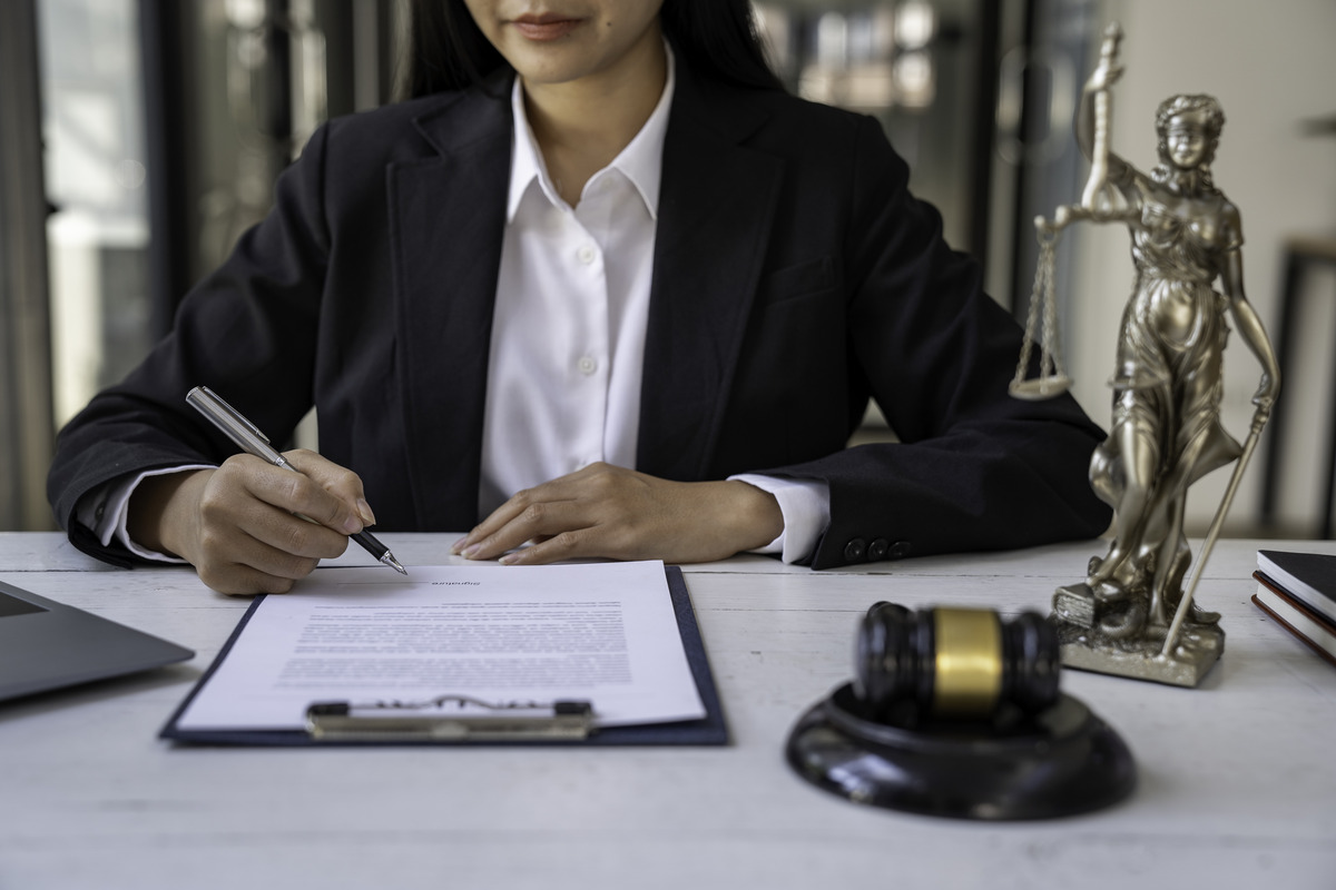 Abogada profesional firmando documentos legales en una oficina con estatua de la justicia y un mazo