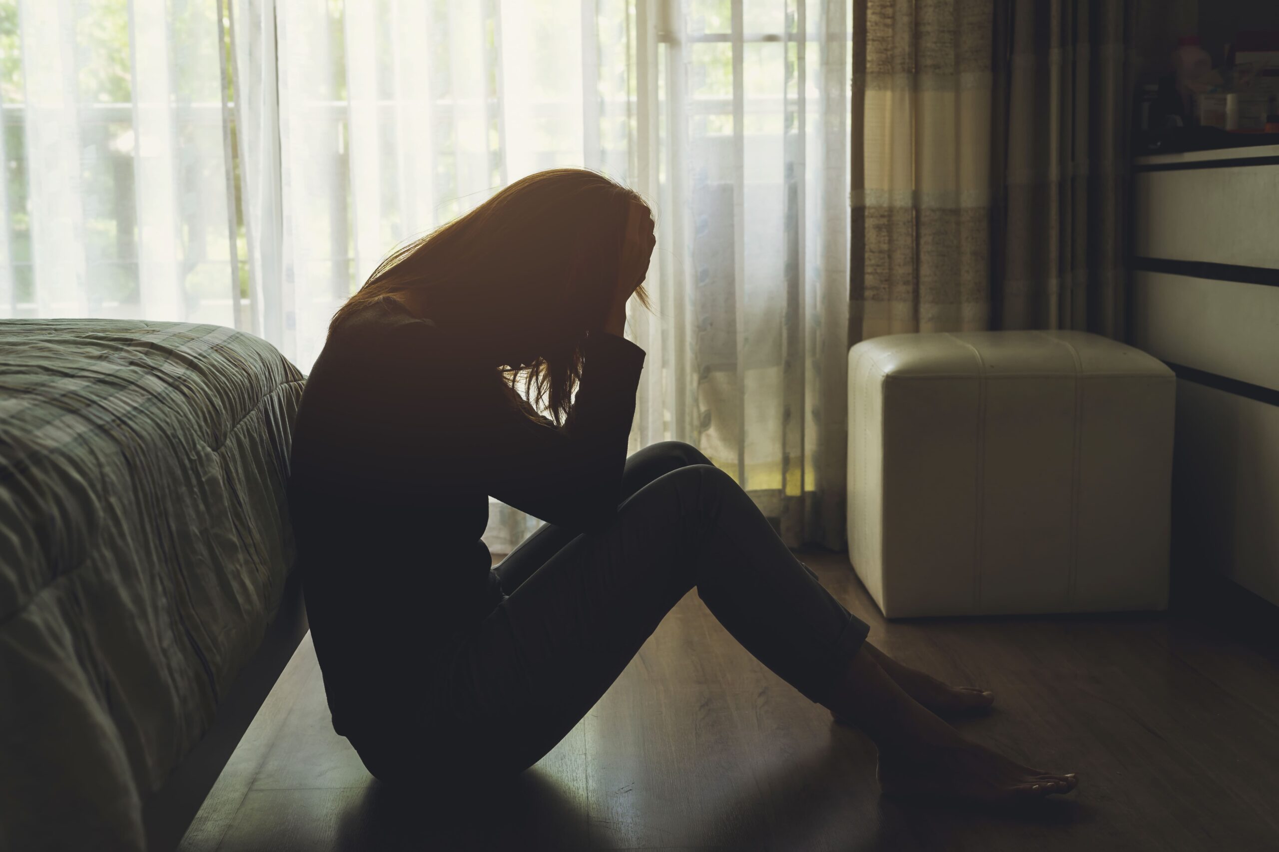 Mujer sentada sola en el piso de una habitación con la cabeza entre las manos cerca de una ventana, representando angustia emocional, problemas de salud mental, depresión, recuperación de traumas y el impacto de los desafíos personales.