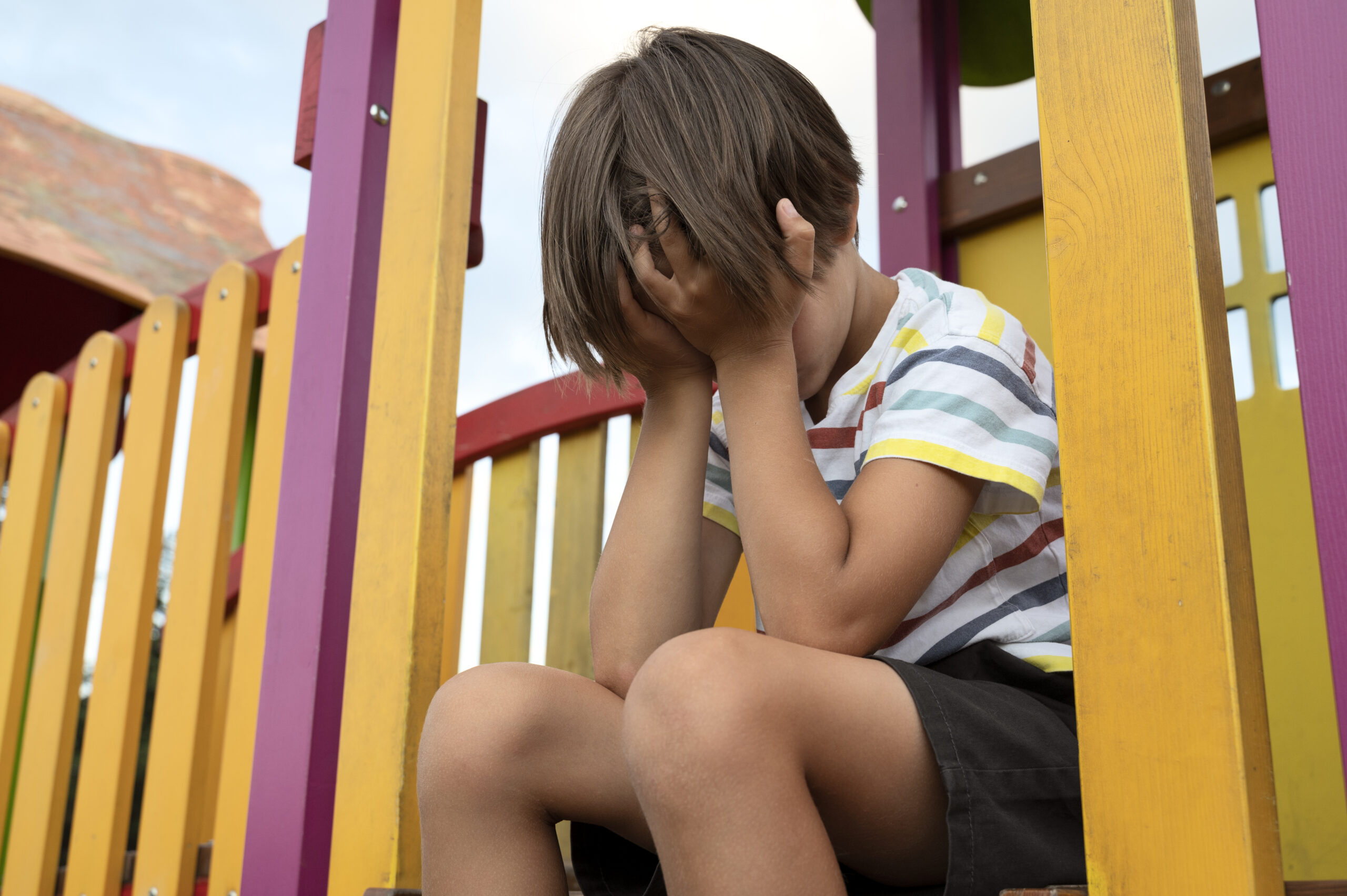 Niño triste sentado solo en un área de juegos con el rostro cubierto por las manos, representando angustia emocional, ansiedad infantil, desafíos familiares, salud mental y bienestar infantil