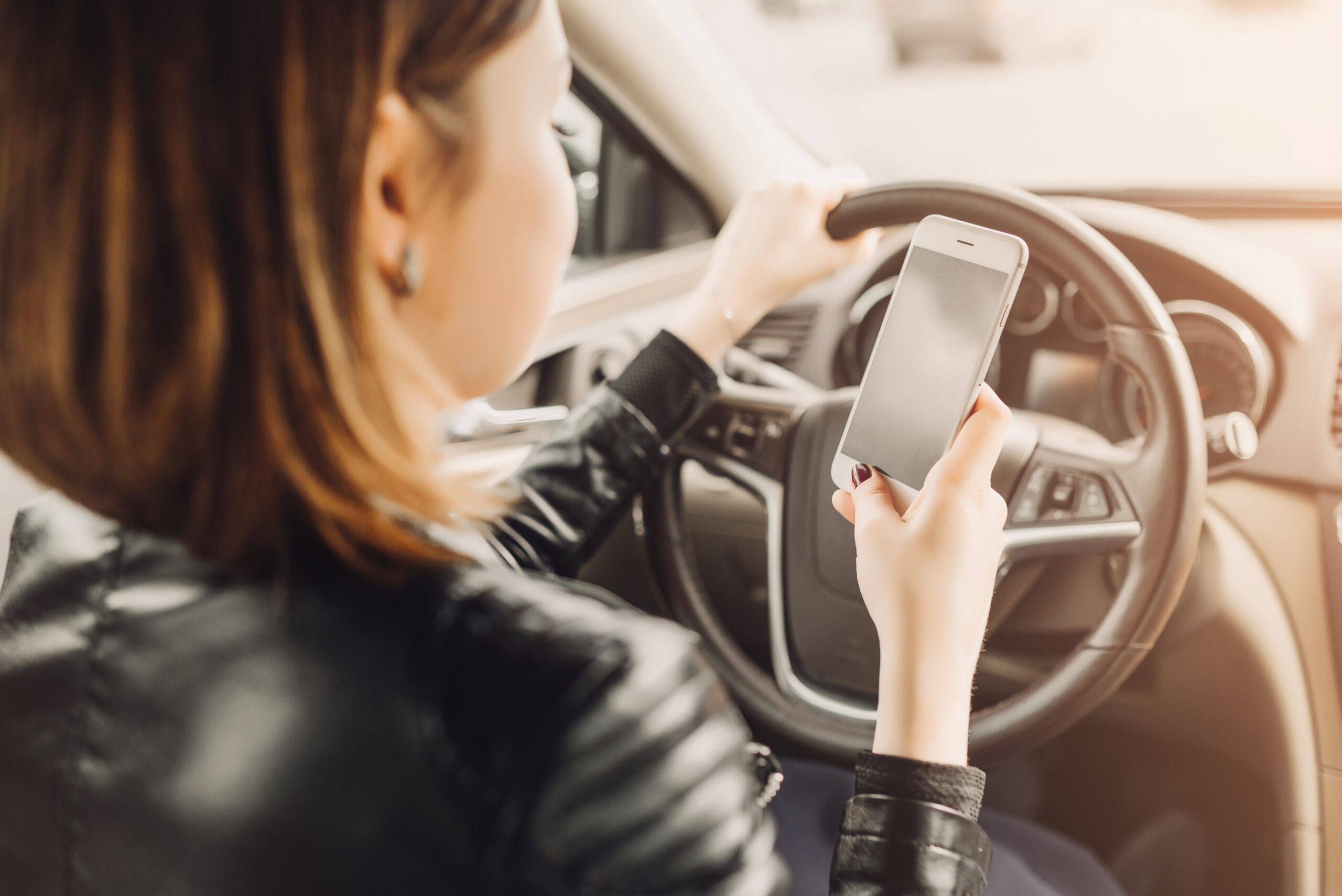 Conductor sosteniendo un teléfono inteligente mientras maneja, ilustrando la conducción distraída, enviar mensajes de texto al conducir, el uso del celular al volante, los hábitos de conducción inseguros y el mayor riesgo de accidentes de tránsito