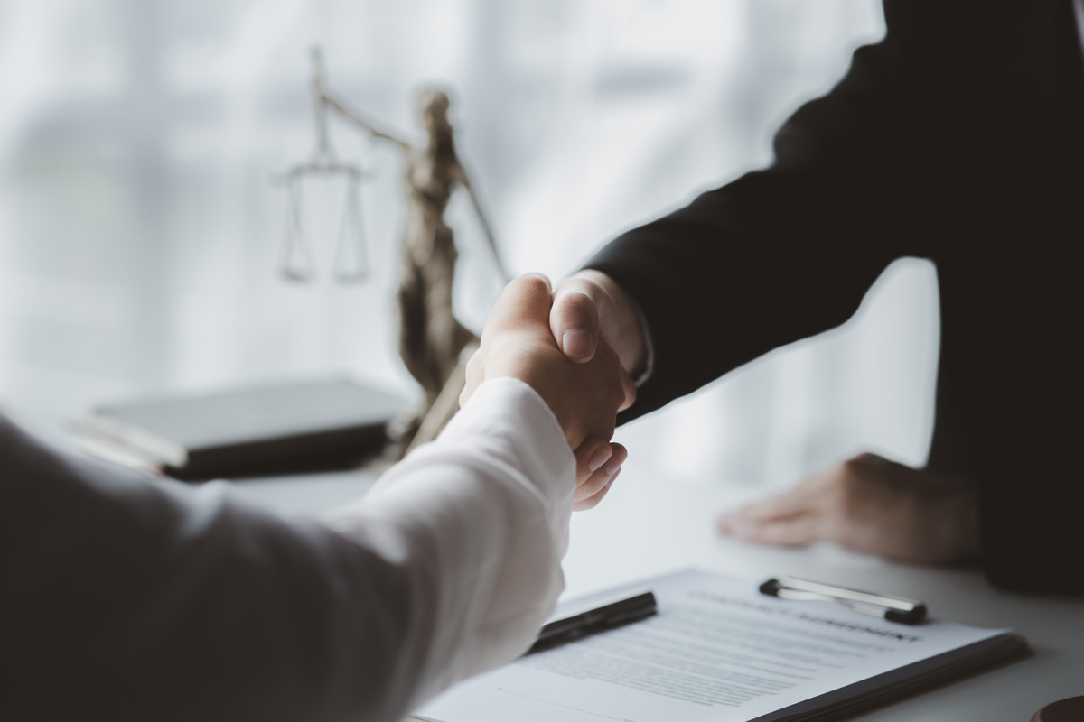 Abogado y cliente estrechando las manos sobre documentos legales con la Dama de la Justicia al fondo, representando una consulta legal, la relación abogado-cliente, la resolución de casos, la representación legal de confianza y resultados exitosos